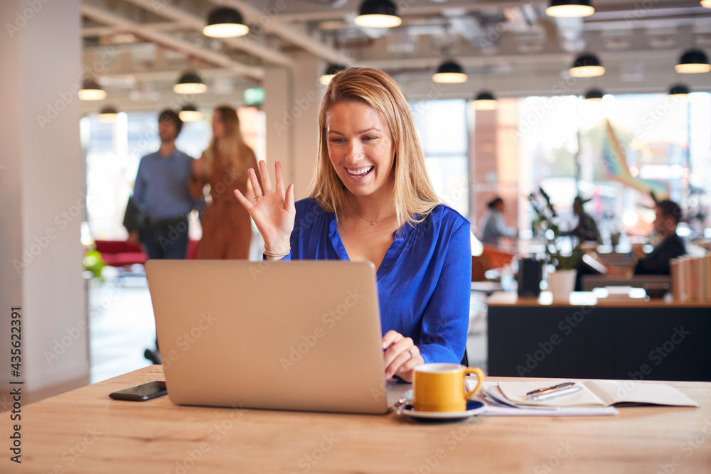 Young Businesswoman Sitting At Desk On Video Call In Modern Open Plan Office Young Businesswoman Sitting At Desk On Video Call In Modern Open Plan Office
