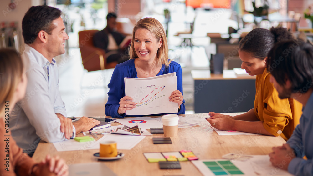 Businesswoman Giving Presentation To Colleagues Sitting Around Table In Modern Open Plan Office