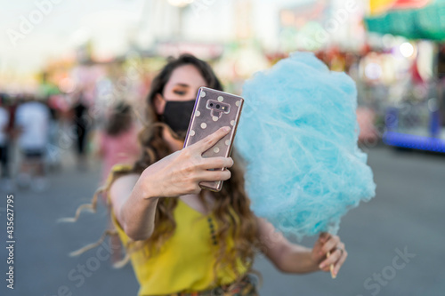 Chica joven con mascarilla por covid 19 tomando un algodón de azucar azul y tomando selfies con el movil en una zona de atracciones de una feria photo