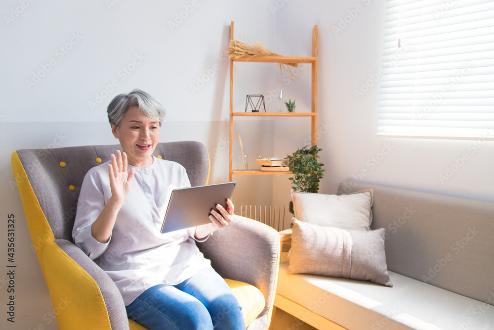 Smiling Asian Senior woman sitting in sofa making video call on digital tablet. Woman waving hand while talking with people via wireless internet technology. Smiling Asian Senior woman sitting in sofa making video call on digital tablet. Woman waving hand while talking with people via wireless internet technology.
