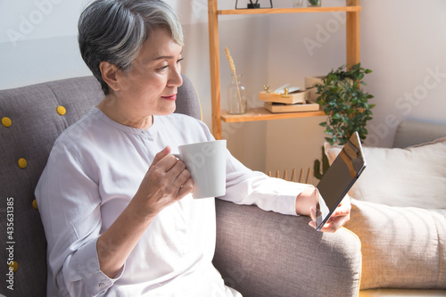 Joyful senior woman drinking coffee and looking at her digital tablet in the morning at home.