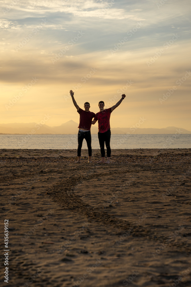 Two silhouettes of men on the beach. In a sunset with a calm sea.Friends holding each other by the back and raising an arm in joy.