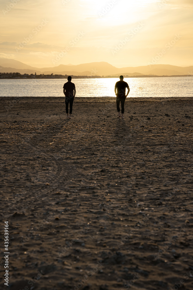 Two men walking towards the sea water. At sunset. They walk apart.