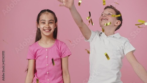 Studio portrait of happy little boy and girl throwing golden glitter up and enjoying rain, dancing over pink background photo