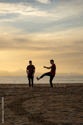 Two silhouettes of men on the beach. In a sunset with a calm sea.One of them is kicking the air and the other has his hands in his pockets.