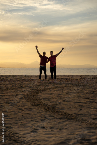 Two silhouettes of men on the beach. In a sunset with a calm sea.Friends holding each other by the back and raising an arm in joy.