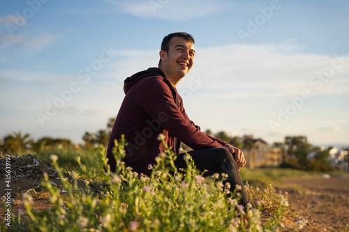 Happy man sitting on the beach smiling. He is wearing a red sweatshirt and black trousers. Caucasian © J.Poquet