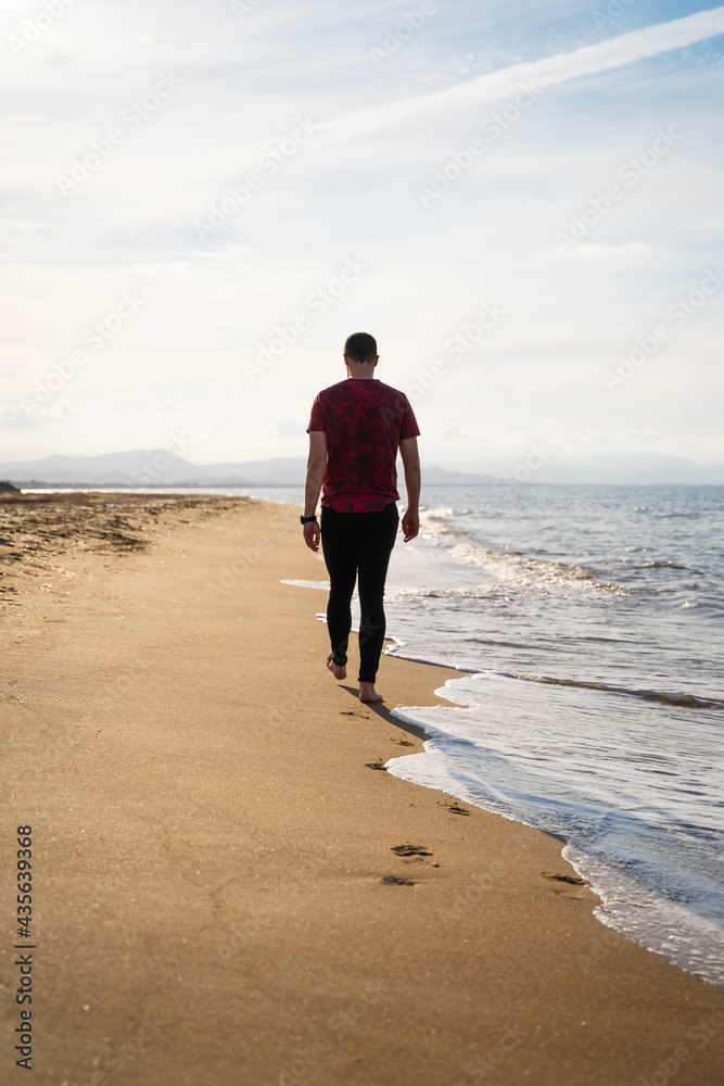 A Caucasian man is walking along the seashore. It is a sunny day and the sea is calm. He is wearing a red short-sleeved T-shirt and black jeans. He is alone on the beach. Les Marines, Denia. A Caucasian man is walking along the seashore. It is a sunny day and the sea is calm. He is wearing a red short-sleeved T-shirt and black jeans. He is alone on the beach. Les Marines, Denia.