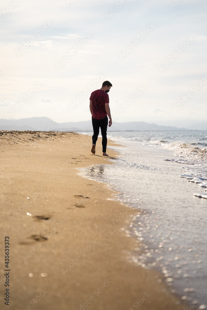 A Caucasian man is walking along the seashore while watching the waves. It is a sunny day and the sea is calm. He is wearing a red short-sleeved T-shirt and black jeans. He is alone on the beach.