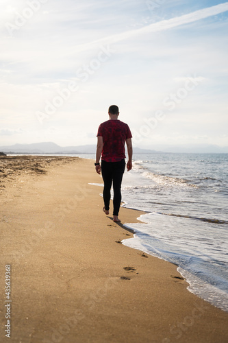 A Caucasian man is walking along the seashore. It is a sunny day and the sea is calm. He is wearing a red short-sleeved T-shirt and black jeans. He is alone on the beach. Les Marines, Denia.