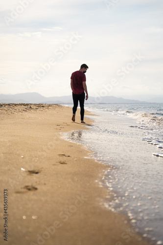 A Caucasian man is walking along the seashore while watching the waves. It is a sunny day and the sea is calm. He is wearing a red short-sleeved T-shirt and black jeans. He is alone on the beach.