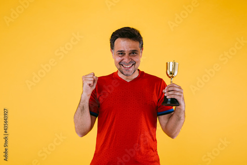 A soccer fan in a red jersey, he clenches his fist and holds a winner's trophy.