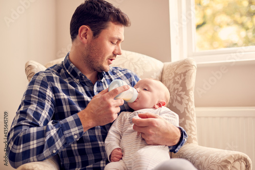 Father Feeding Baby Son With Bottle Sitting On Chair In Lounge At Home Together