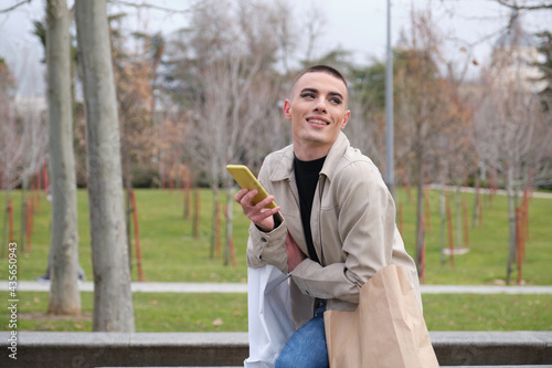 Young man wearing make up holding shopping bags and smartphone laughing sitting in a park. Queer person.