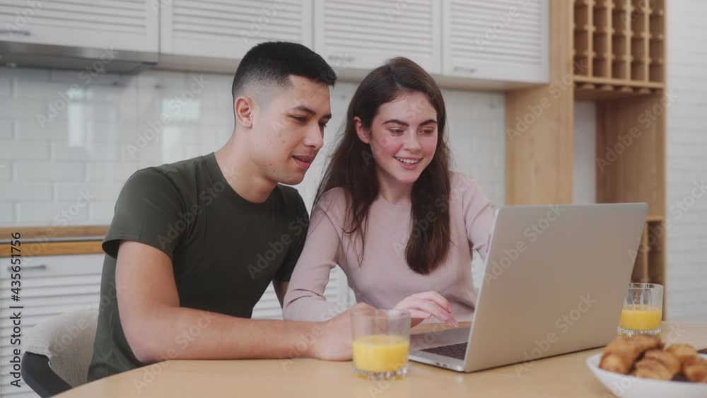 The happy couple looking at the laptop screen and having breakfast while sitting at the table in the kitchen The happy couple looking at the laptop screen and having breakfast while sitting at the table in the kitchen
