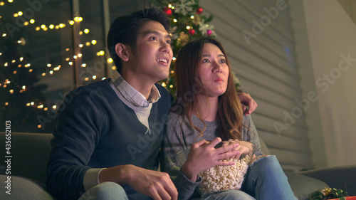 Stock video.A young Asian couple sits on a sofa in a decorated room to celebrate Christmas and New Year. Man and girlfriend eating popcorn and enjoying to watching TV show.