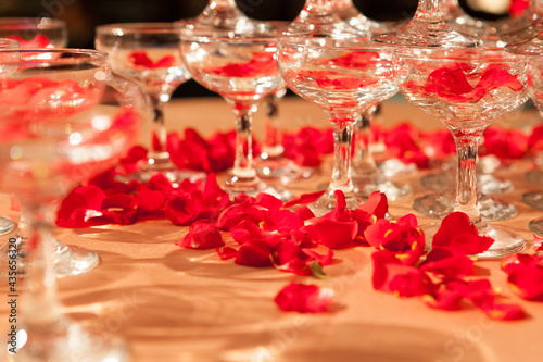Red rose petals are spilled in and around the red wine goblet on the golden banquet table, accompanied by the reflection of the light photo