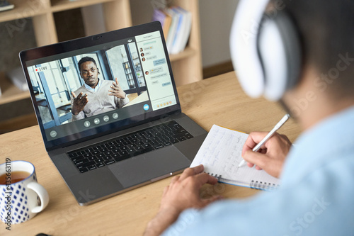 Latin indian adult student wearing headset having virtual meeting online call training educational webinar chatting with black teacher at home office using laptop. Over shoulder view.