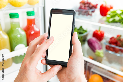 Man using smart phone, buying food. Close-up hand with phone. Grocery shopping online. Empty white screen. Fresh vegetables on fridge shelves. Healthy lifestyle and food delivery.
