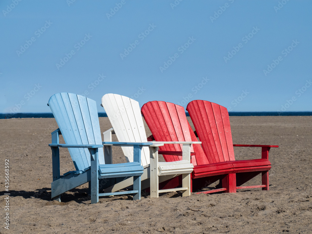 Red, white and blue Adirondack chairs on a sandy beach on a sunny day
