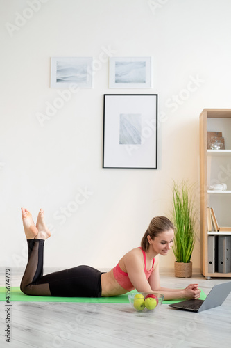 Fitness blog. Sportive woman. Online communication. Happy athletic lady in sportswear resting yoga mat looking laptop with bowl fresh apples in light room interior.