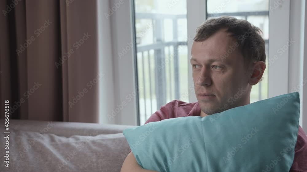 Portrait of a young man immersed in his thoughts sits on a sofa clutching a pillow to himself Portrait of a young man immersed in his thoughts sits on a sofa clutching a pillow to himself