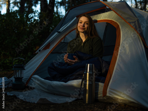 In the photo it is late evening. Dusk. A young woman is sitting in a tent. There is a thermos next to her .. The woman is holding a cup. There is a dense dark forest behind the tent.