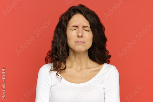 Closeup portrait of sad depressed woman with closed eyes suffering from anxiety, depression or headache. Upset unhappy millennial female standing over red wall. Tired girl in emotion of sorrow or pain