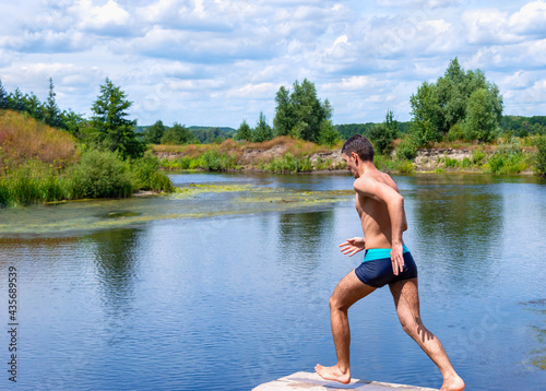 Teenager running on a wooden bridge, sprints to jump into the river. Sports guy jumping into the water on a sunny day. Beautiful landscape of a river with green banks. Concept of healthy lifestyle