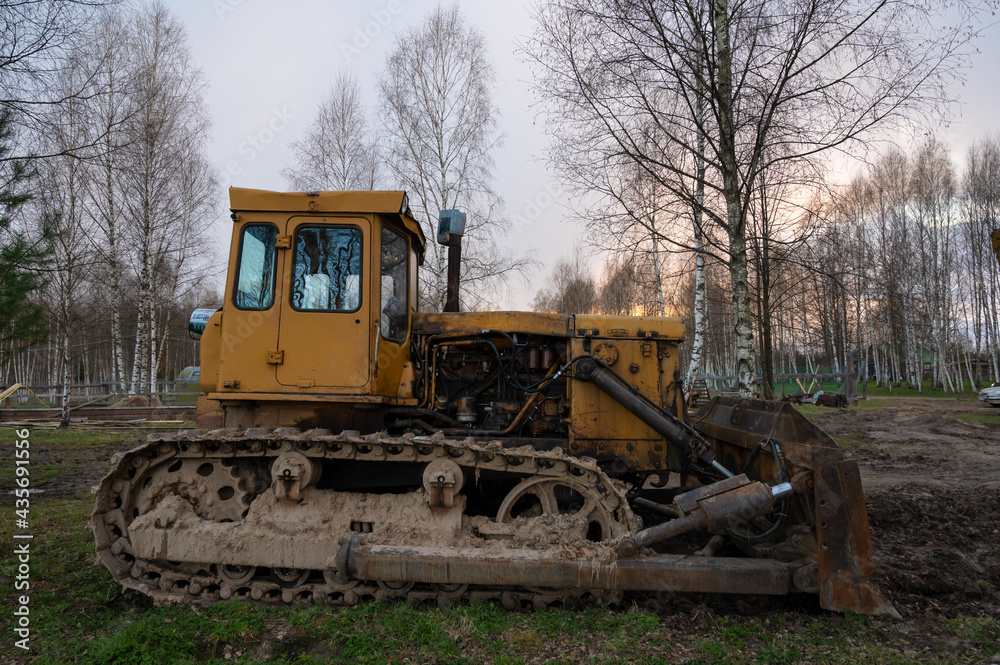 An old rusty yellow tractor among the trees in the open air on a driverless farm. Agricultural machinery An old rusty yellow tractor among the trees in the open air on a driverless farm. Agricultural machinery