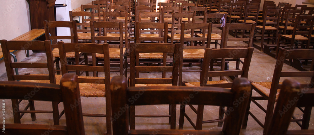 View on isolated old brown wood chairs with backrest in a row inside catholic empty church illuminated by natural sun light (focus on backrest of chair in center))