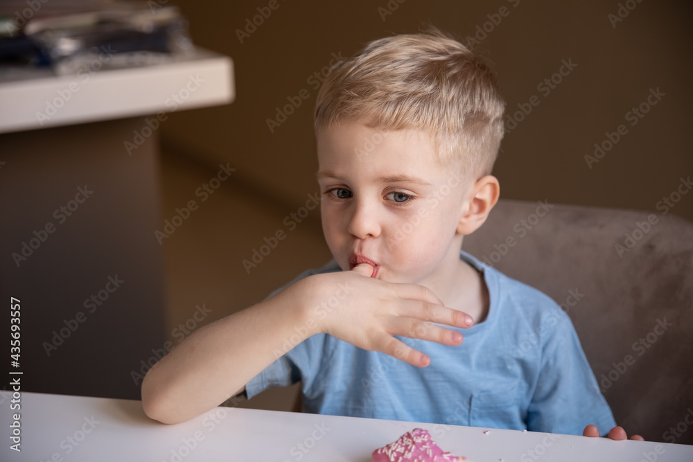 The concept is useless food. A little boy eats a pink doughnut at home and licks his fingers. He likes the doughnut. The concept is useless food. A little boy eats a pink doughnut at home and licks his fingers. He likes the doughnut.