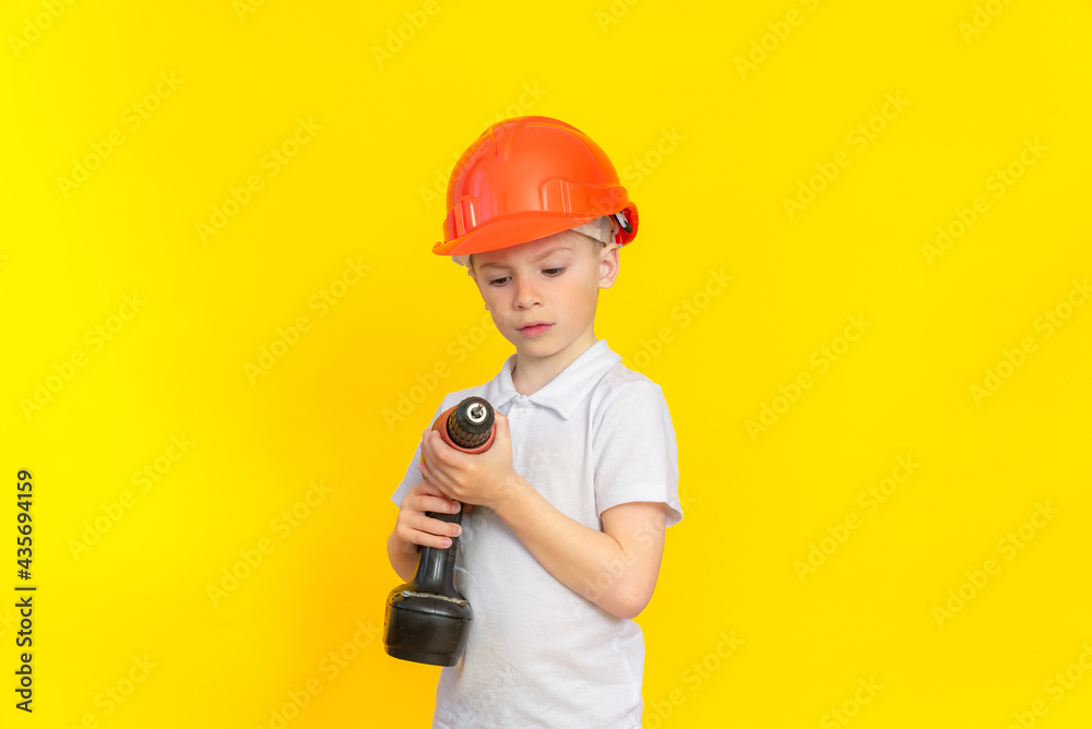 Building craft training. A boy in a kaskuke stands on a yellow background and looks attentively at a construction electric tool. The profession is a builder, the development of children.