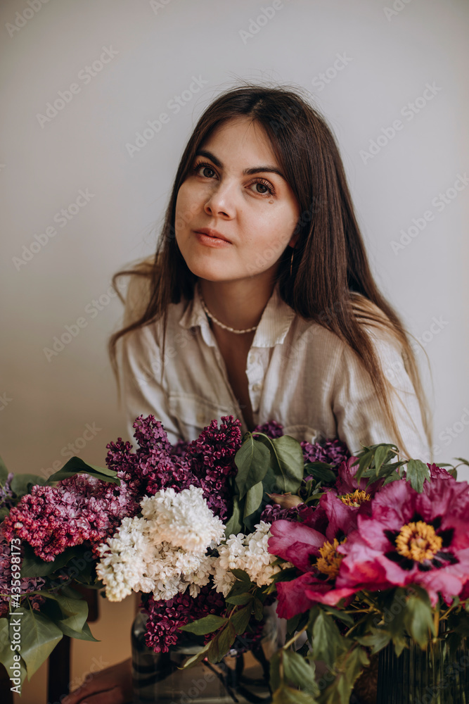 young beautiful woman is sitting near her bouquets of flowers. the girl was presented with bouquets of lilacs and peonies. brunette girl with brown eyes in a white shirt near flowers. young beautiful woman is sitting near her bouquets of flowers. the girl was presented with bouquets of lilacs and peonies. brunette girl with brown eyes in a white shirt near flowers.
