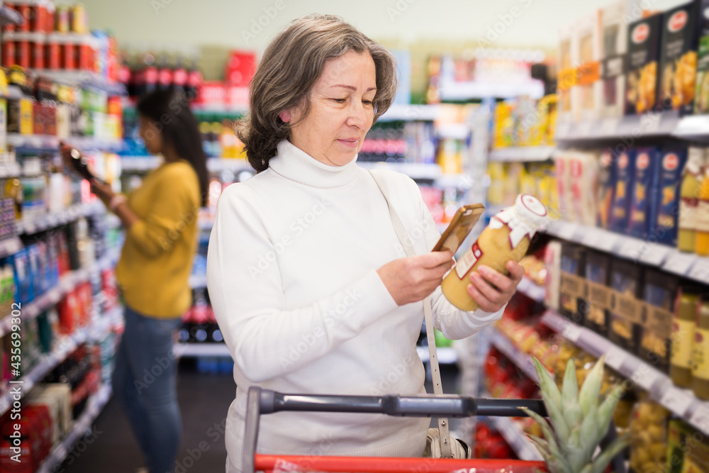 Positive interested elderly woman scanning barcode on bottle of fruit juice with her smartphone, checking product information. Concept of modern technologies for convenience of buyers