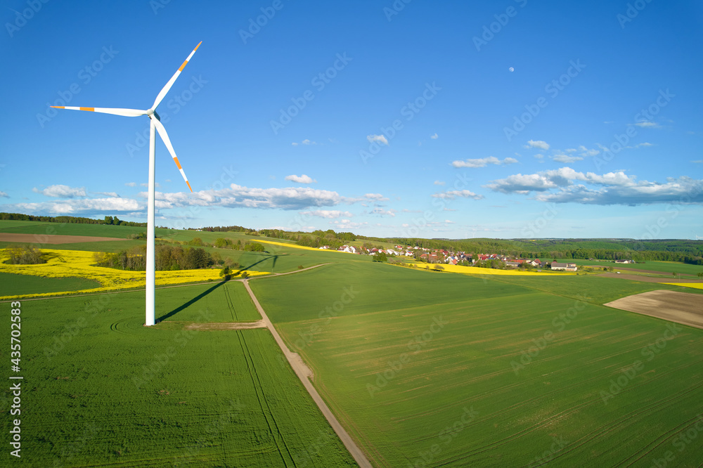 Obraz premium 1 wind turbine on green farmland. Small village in the background. Blue sky with small moon. Aerial view. Front view.