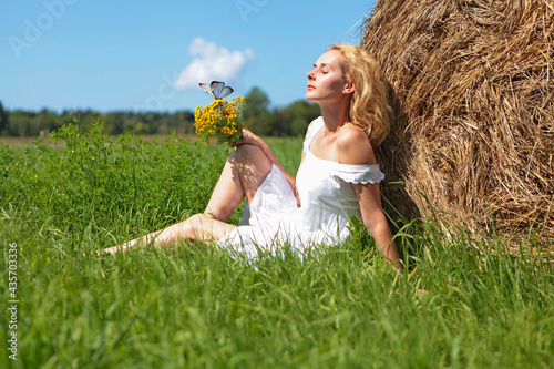 Blond young beautiful woman sitting in white dress near hay ball at the green field with yellow flowers and blue butterfly. Summer vacation and joy concept. photo