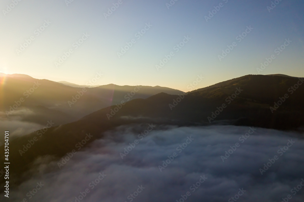 Aerial view of vibrant sunrise over white dense fog with distant dark silhouettes of mountain hills on horizon.