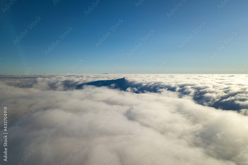 Aerial view from above of white puffy clouds and distant mountain top in bright sunny day.