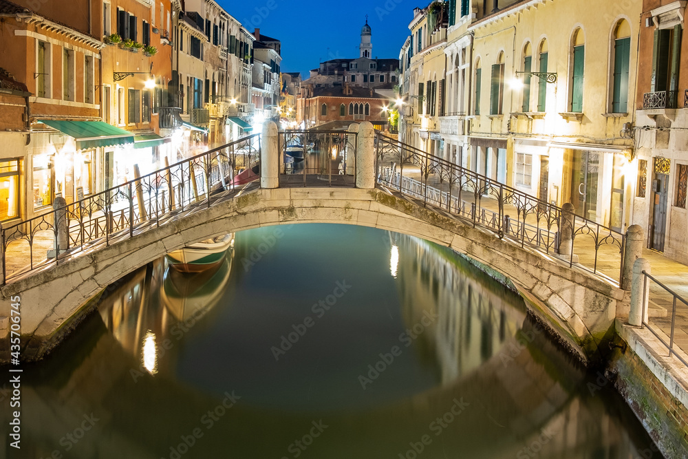 Night view of illuminated old architecture, floating boats and light reflections in canals water in Venice, Italy.