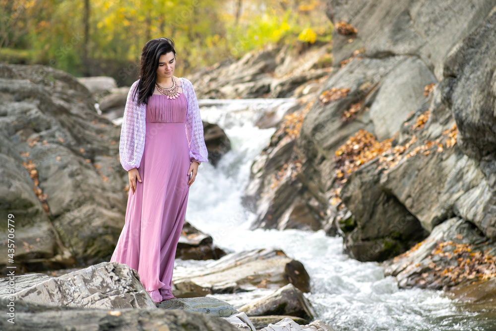 Young pretty woman in long pink fashionable dress standing near small river with fast moving water.