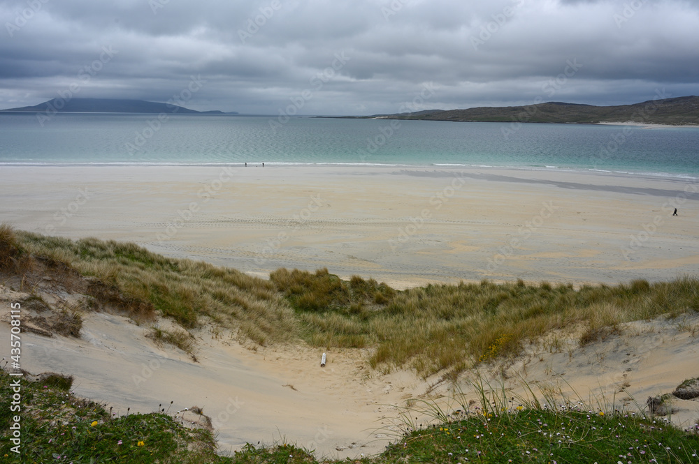 Luskentyre Beach, Isle of Harris, Scotland. Moody day with cloudy sky. Showing sandy beach, turquoise sea, sand dunes with grass and flowers. Unidentifiable people silhouetted on beach.