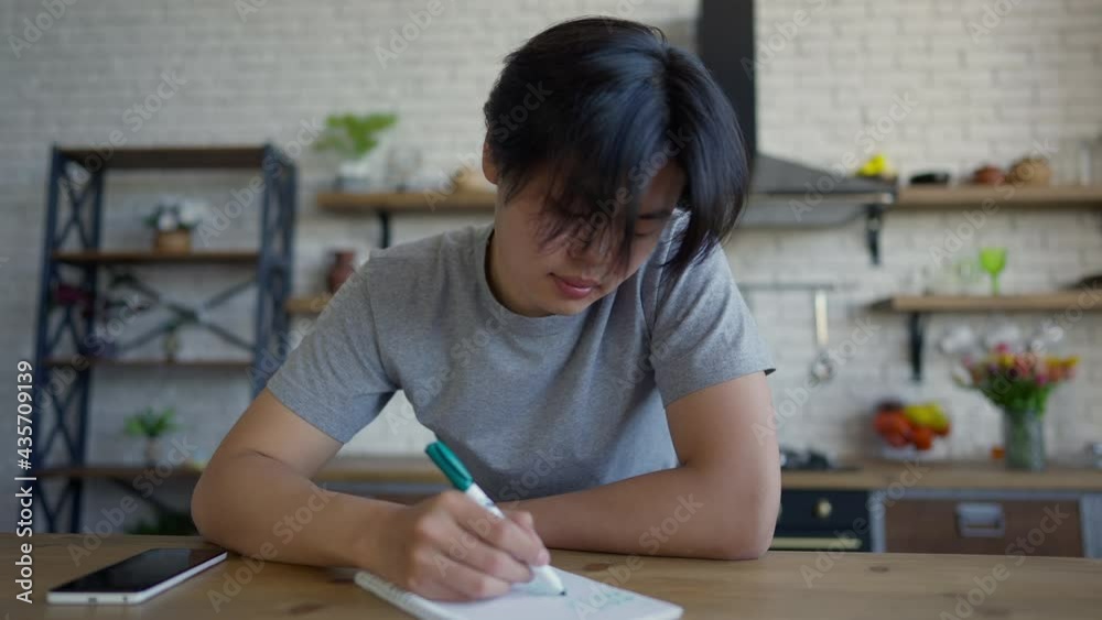 Smiling concentrated Asian man writing with pen in sketchpad sitting at kitchen table indoors. Front view portrait of confident entrepreneur handwriting startup ideas in home office