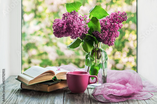 Morning coffee. A cup of coffee or tea, a bouquet of purple lilacs and an open book on a wooden table with a view out the window. Still life concept, blurred background.