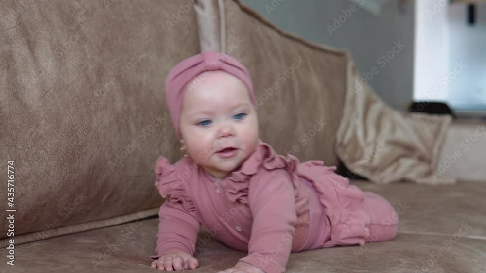 A baby girl with blond hair and blue eyes in pink clothes lies on her tummy and swings in different directions. Baby on a light beige sofa A baby girl with blond hair and blue eyes in pink clothes lies on her tummy and swings in different directions. Baby on a light beige sofa