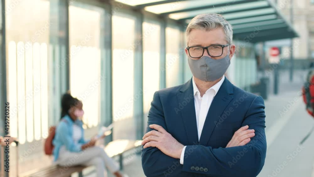 Close up of Caucasian handsome happy middle-aged man in suit standing in city looking at camera at bus station with many people. Male in mask and glasses outdoors, coronavirus pandemic Close up of Caucasian handsome happy middle-aged man in suit standing in city looking at camera at bus station with many people. Male in mask and glasses outdoors, coronavirus pandemic