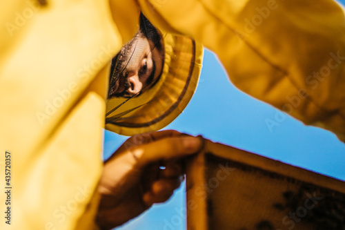 Close portrait shot from ground of a young beekeper with a beard in a yellow protecting suit working on his apiary on a summer day. photo