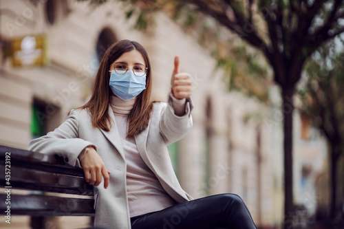 Girl with face mask on sitting on a bench outside during corona virus, looking at camera and showing thumbs up. Millennials during lockdown. © dusanpetkovic1