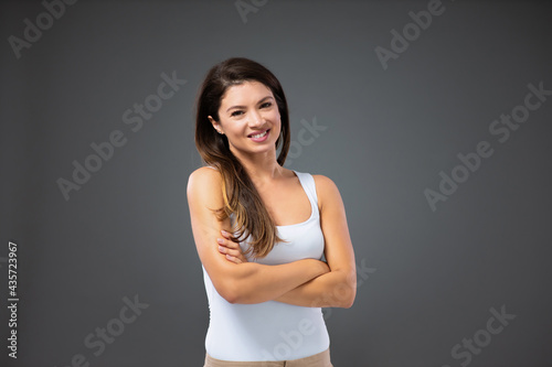 Portrait of a young woman with a pleasant smile, long dark hair, and healthy skin wears a white T-shirt. Female crossed her arms over her chest and stands on a gray background being in a good mood