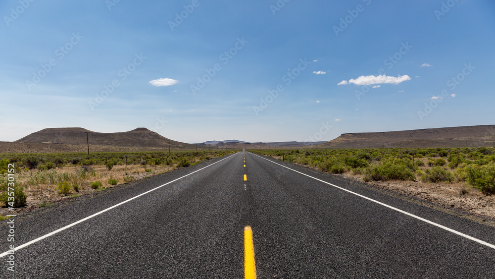 Straight asphalt road with dashed yellow strip line running between hills into horizon. A road in a high desert of South Oregon